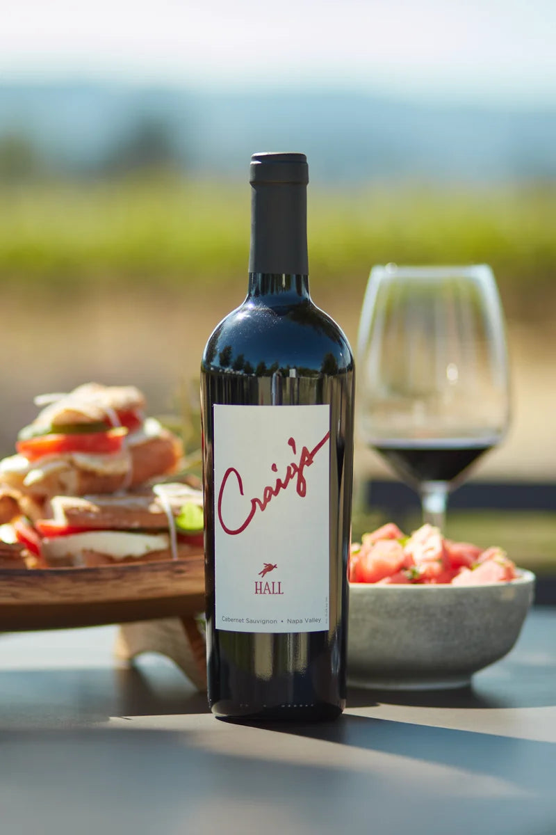 Photo of a bottle of 2020 HALL Craig's Cabernet Sauvignon on a picnic table with a platter of sandwiches, a summer salad, and glass of red wine in the background.