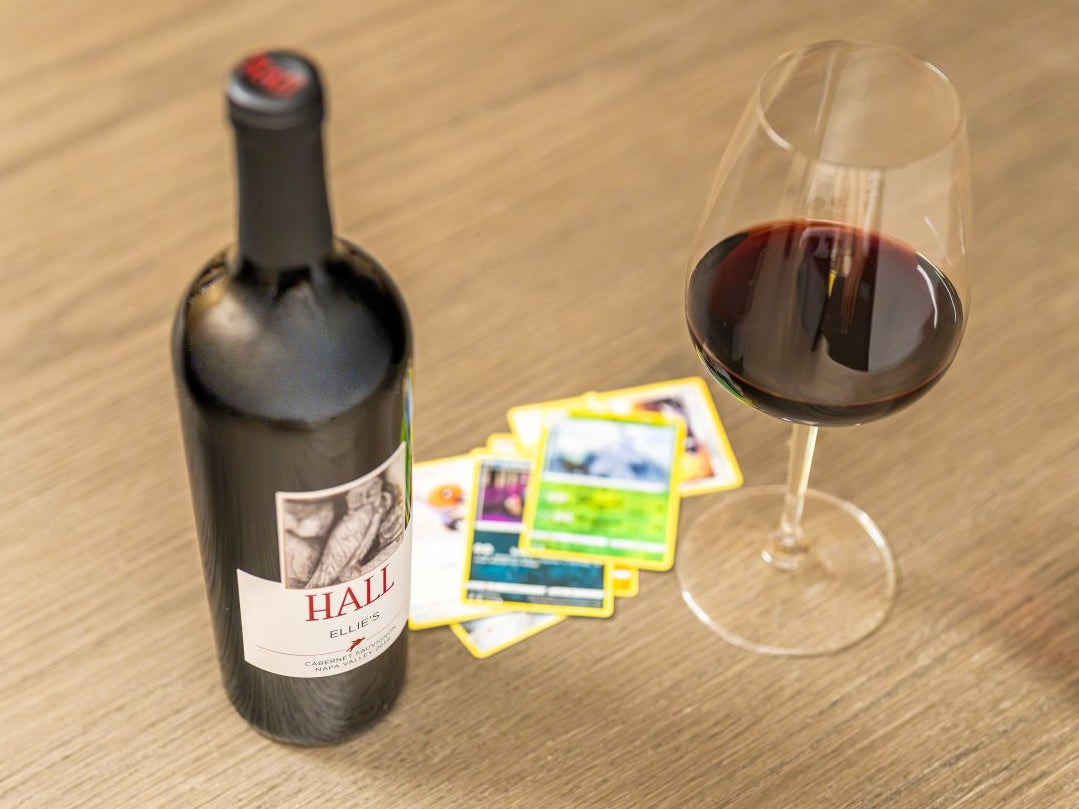 A bottle of wine and a glass of red wine on a wooden table, with a few colorful cards fanned out between them. The bottle label shows the brand HALL.