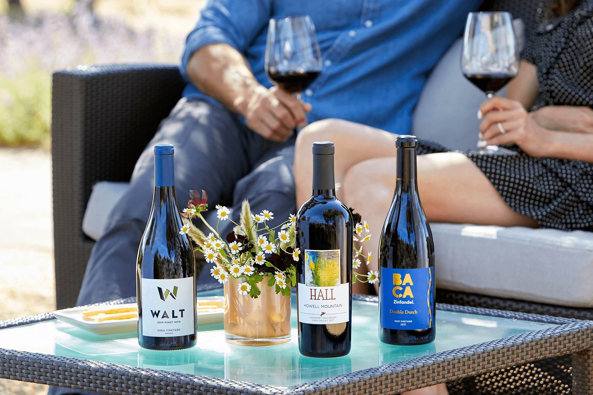 Three wine bottles from HALL, WALT and BACA wines on a table at WALT/BACA Healdsburg with people tasting wine in the background.