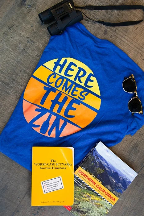 A flat lay image featuring a Blue BACA Wines t-shirt with a sunset design that says "here comes the zin". The image also contains a pair of binoculars, sunglasses and two hiking guide books.