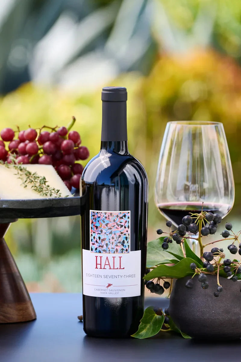 Bottle of 2020 HALL Eighteen Seventy-Three Cabernet Sauvignon beside a cheese and fruit platter and a glass of red wine