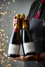 Image of the Michel Foch Brut & Rosé Duo gift set. The image is of a man wearing an apron and holiday tie holding the two bottles of Champagne included in this gift set with twinkling lights in the background.