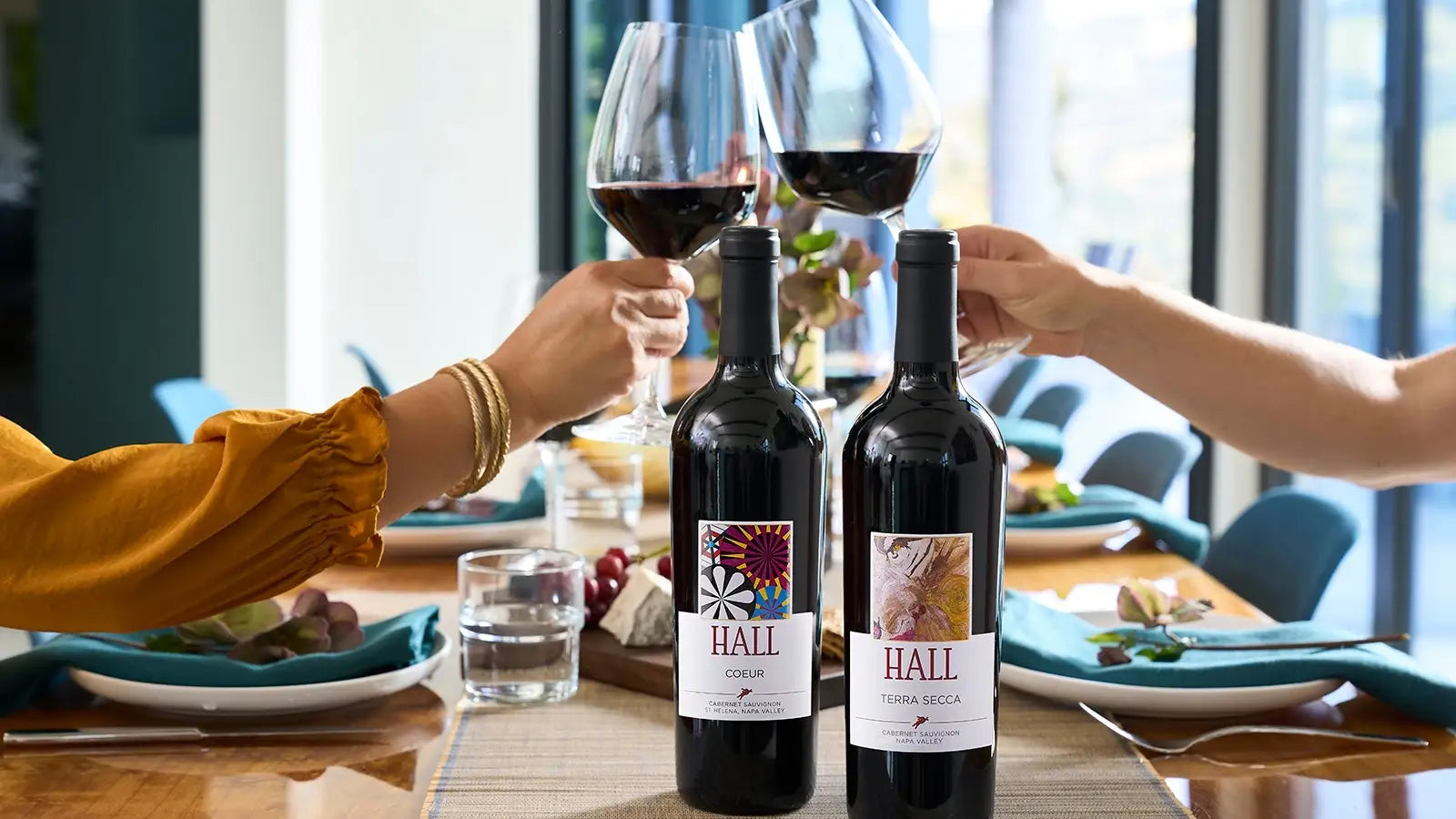 HALL Coeur and Terra Secca Napa Valley Cabernet Sauvignon bottles on a dining table while two people toast with glasses of red wine for the Primary Colors wine club.